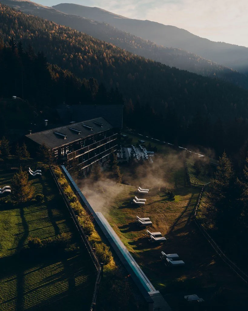A hillside resort surrounded by pine trees, with a large building, a grassy area, outdoor lounge chairs, and mountain landscape in the background during sunset.