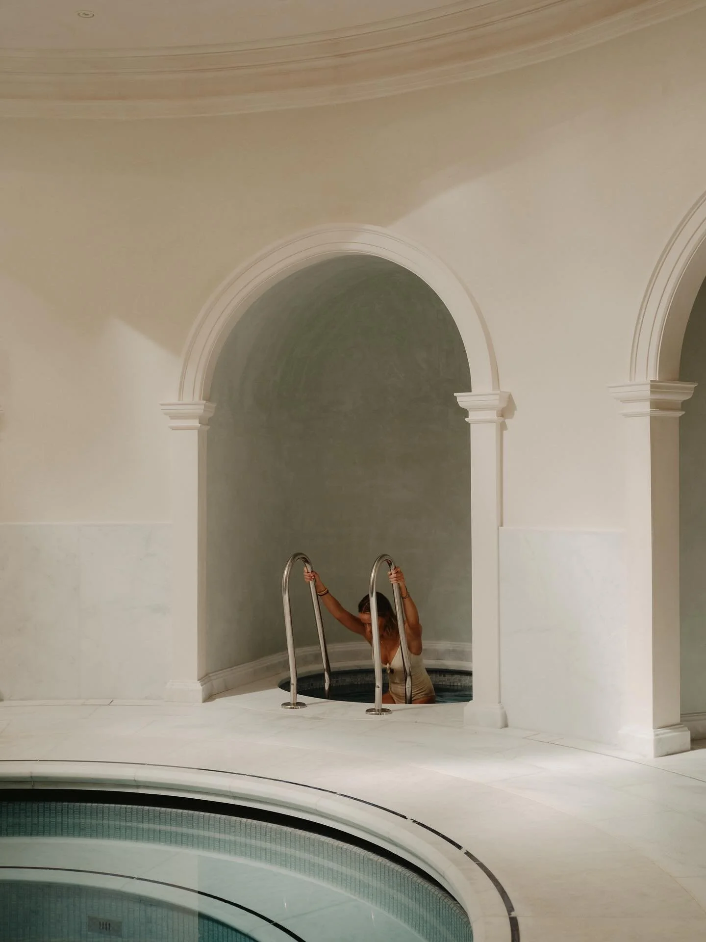 A woman in beige clothing is entering a circular hot tub or pool, holding onto the metal handrails, in a room with beige walls and architectural arches.