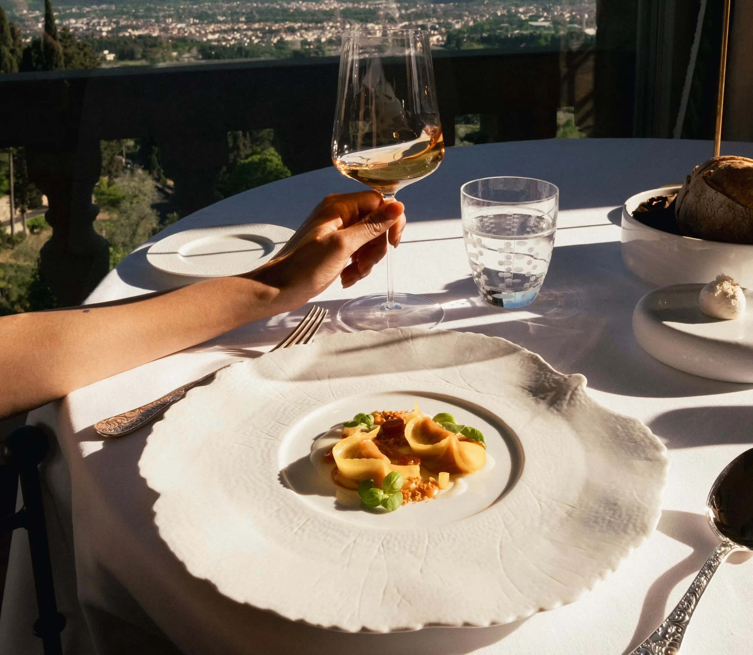 A hand holding a glass of rosé wine at a dining table with a city view in the background. The table has a white plate with pasta, a glass of water, and a bowl of bread.