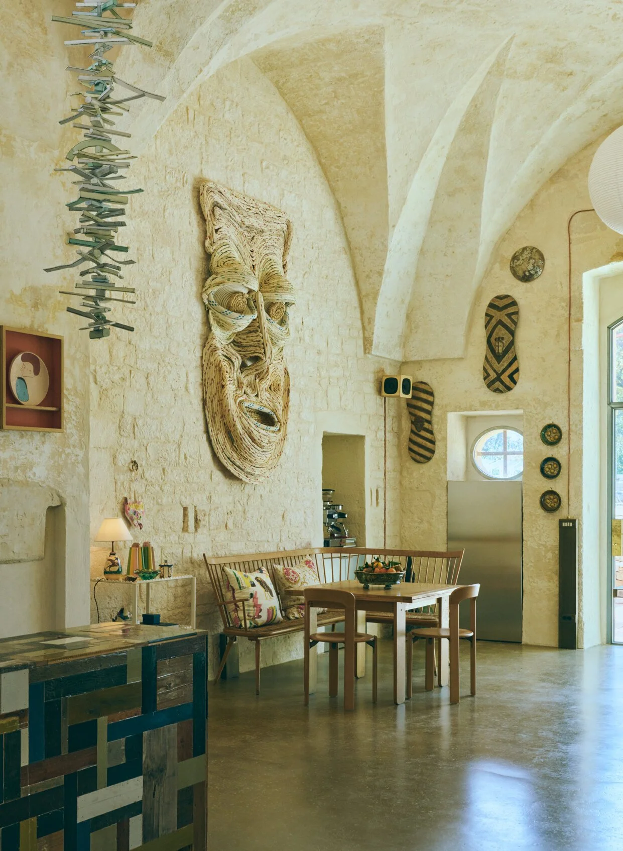 Interior of a room with a high arched ceiling and stone walls, decorated with tribal masks, a large wooden face sculpture, and artwork. There is a round window, a dining table with chairs and cushions, a side table with a lamp, and a small shelf with decorative items.