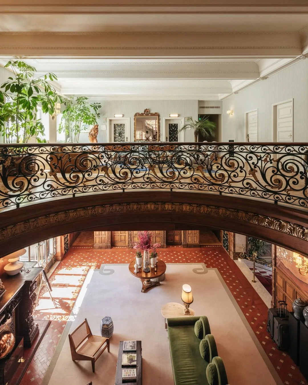 Elegant hotel lobby with ornate wrought iron balcony, lush indoor plants, a large floral arrangement, vintage furniture, and intricate woodwork.