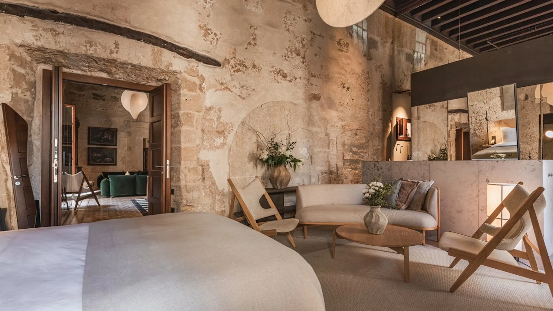 A cozy bedroom with exposed stone walls, a bed with white bedding in the foreground, a seating area with a sofa and chairs, and a view into another room with a green armchair, framed art, and wooden flooring.