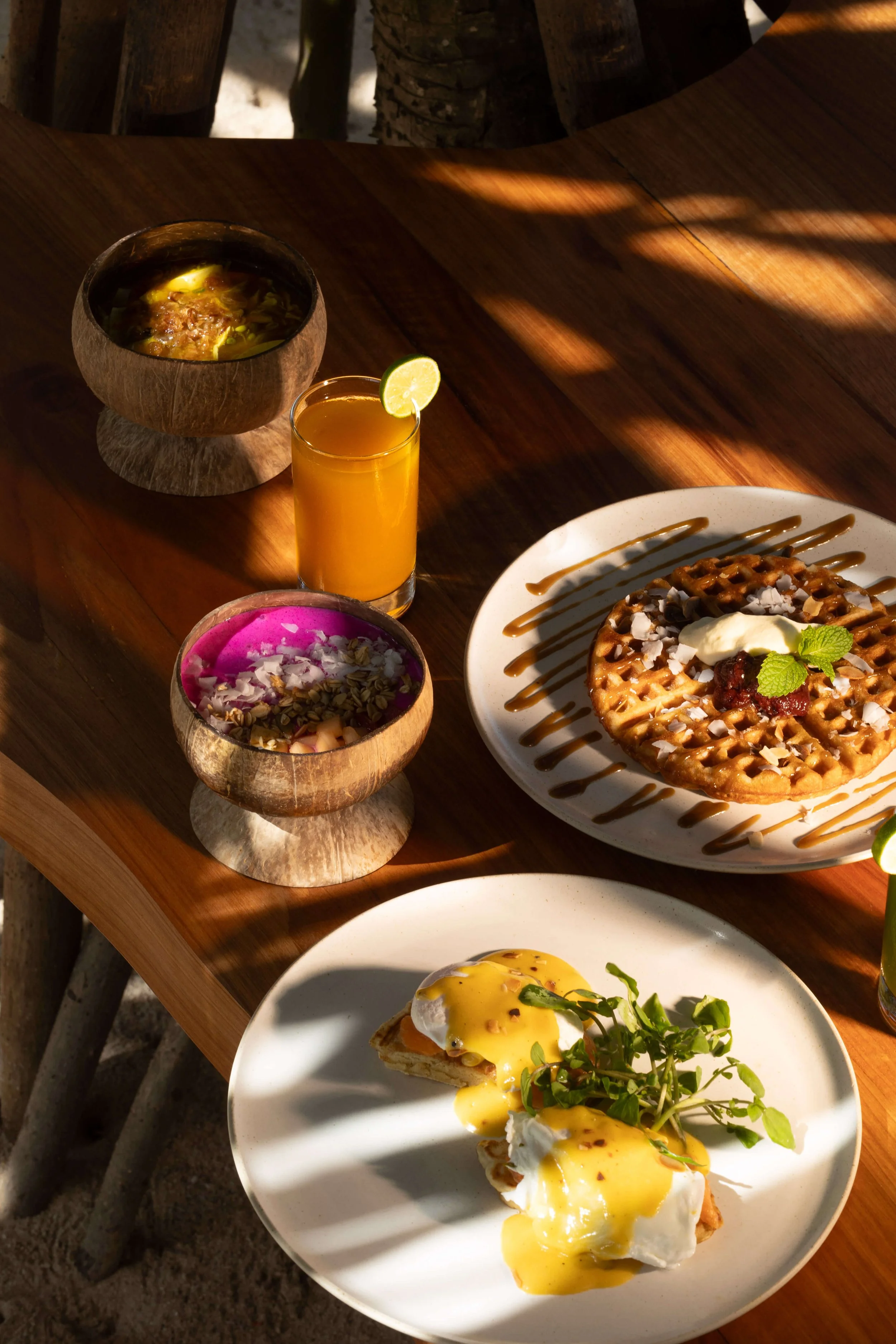 A wooden table holds a wooden bowl of soup, a glass of orange juice with a lime wedge, a purple bowl of yogurt with toppings, and a white plate with waffles topped with ice cream, syrup, and fresh mint, with shadows from sunlight.
