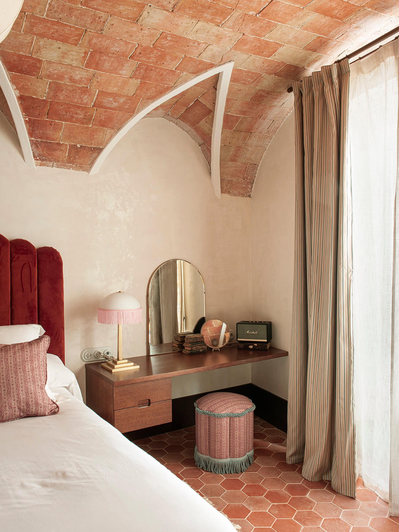 A bedroom corner with a bed, red upholstered headboard, and pink pillow. There is a wooden vanity with an arched mirror, a table lamp with a pink fringe, and vintage items including a small TV and decorative objects. A striped curtain partially covers a window, and the ceiling features exposed brick in an arch shape.