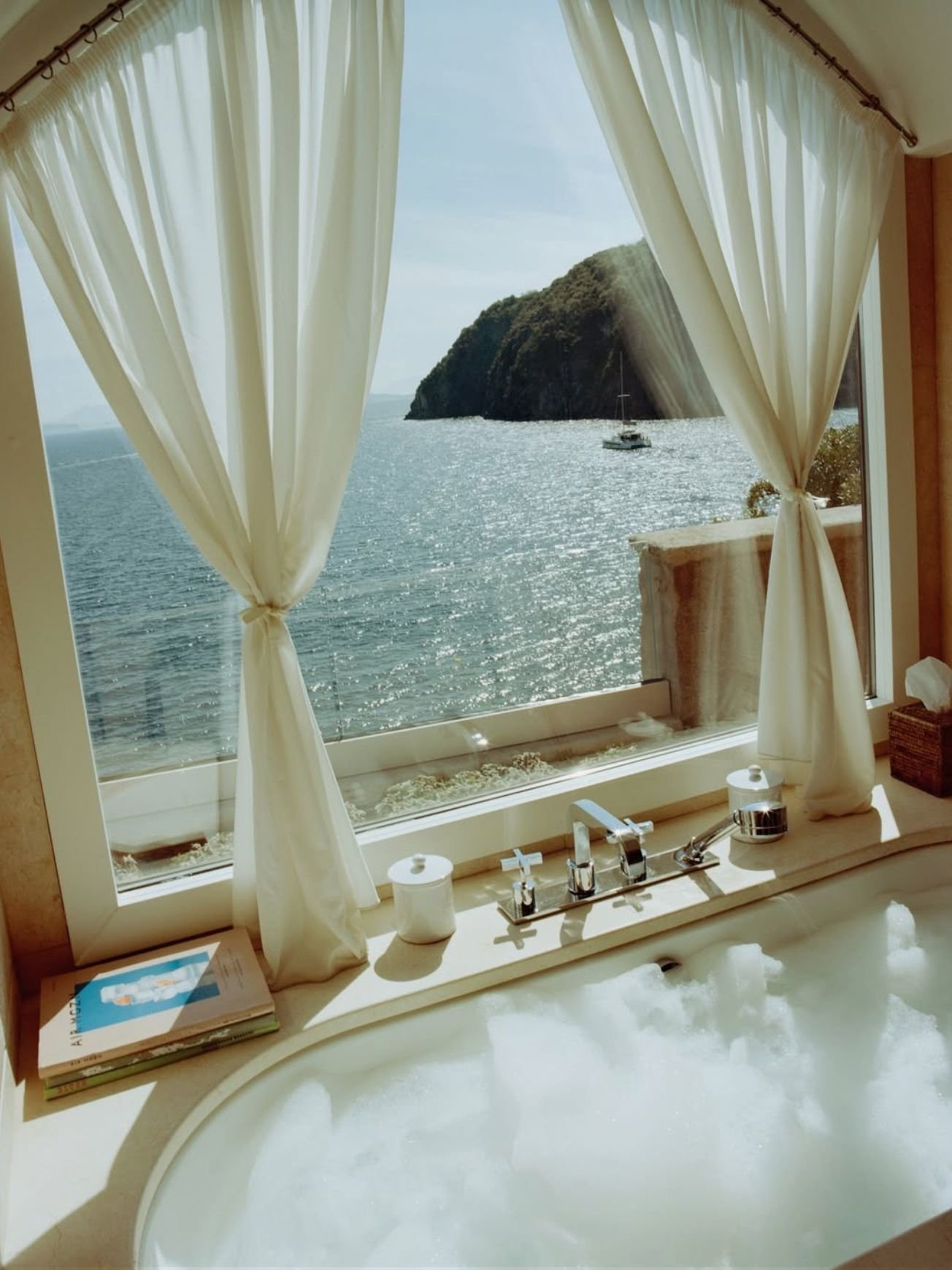 A bathroom with a large window showing a view of the ocean, a boat, and a rocky hill. There are white curtains tied back, a marble countertop, a sink with a faucet, and toiletries. Sunlight streams through the window onto the bathtub filled with bubbles.