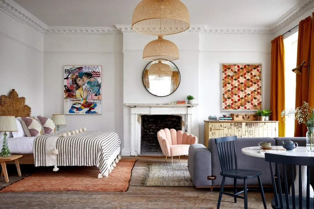 A cozy living room with a bed on the left, a pink velvet armchair near the fireplace, and a gray sofa with a round white table and black chairs. The room has white walls, some colorful artwork, a round mirror, orange curtains, and wooden furniture.