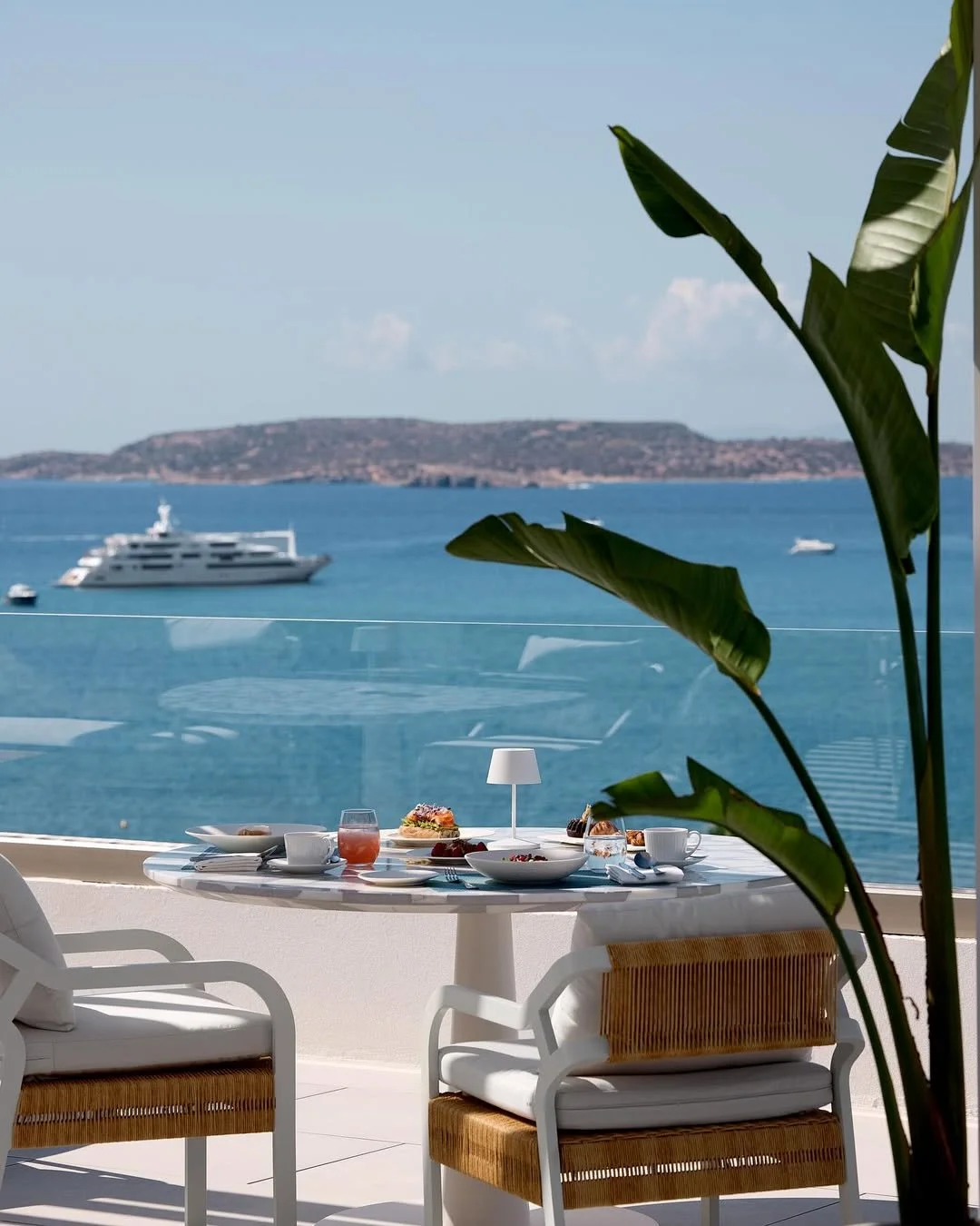 A dining table with breakfast items overlooking a blue sea with boats and a distant landmass, seen through a large window with tropical plants in the foreground.