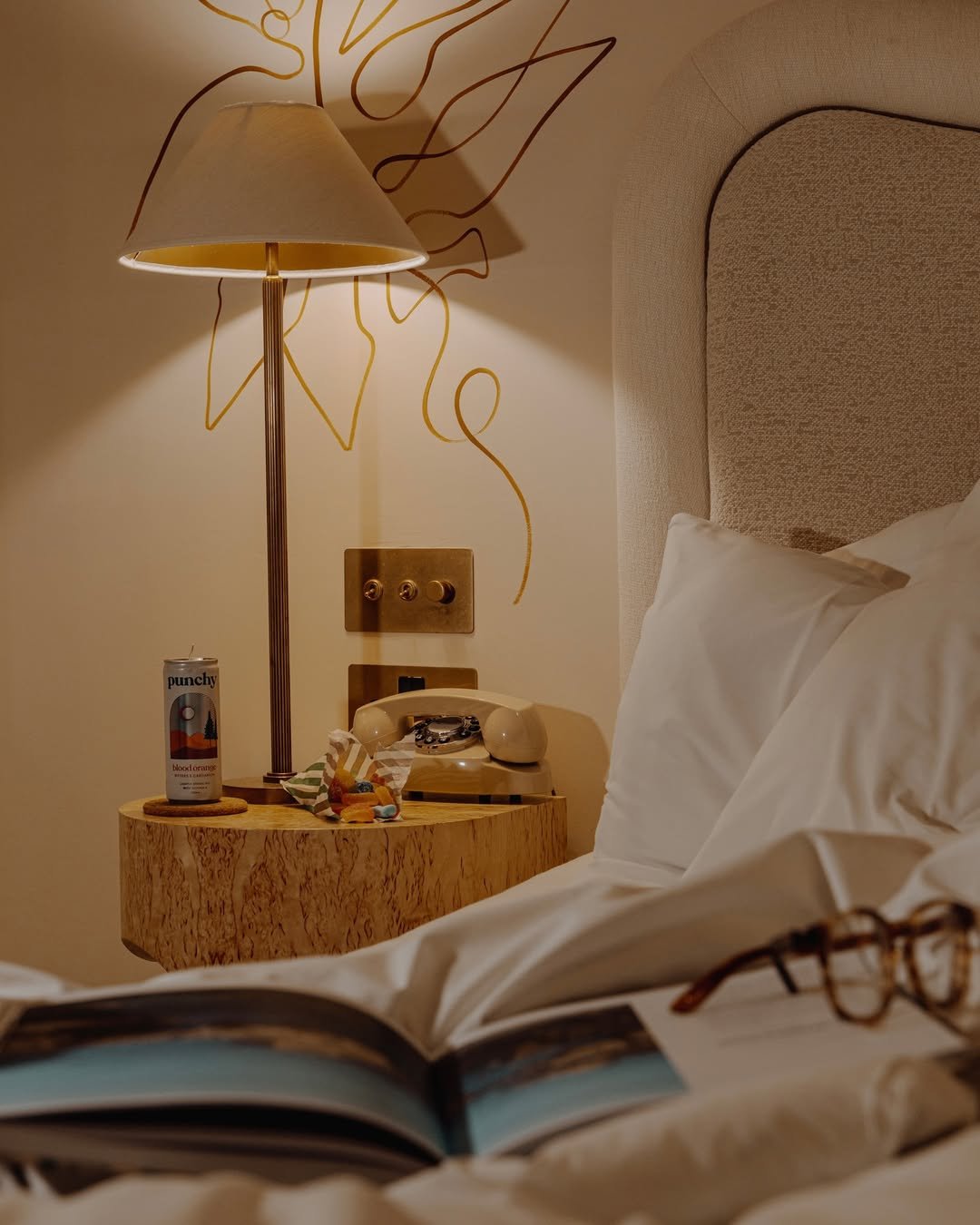 A bedside table with a vintage beige rotary phone, a can of Punchy blood orange drink, candy, and a pair of eyeglasses; a beige bed with pillows; a wall with abstract line art and a lamp casting warm light.