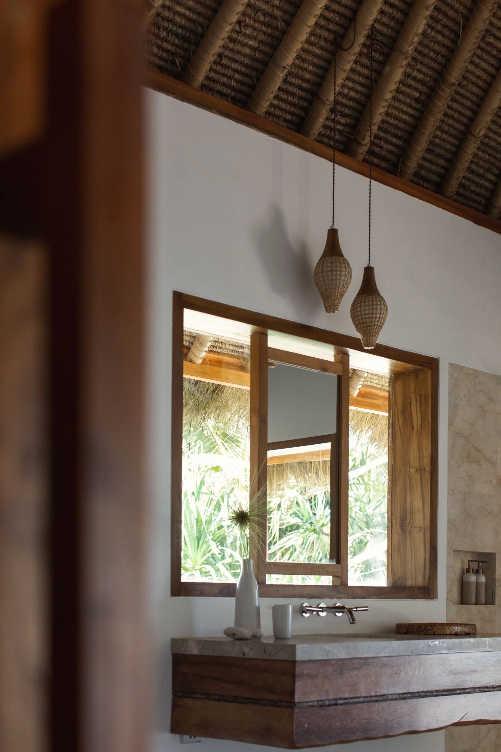 A bathroom vanity with a white countertop, a ceramic vase, and decorative bottles. A large rectangular mirror with a wooden frame reflects green tropical plants outside through a window. Two hanging woven pendant lights are above the vanity.