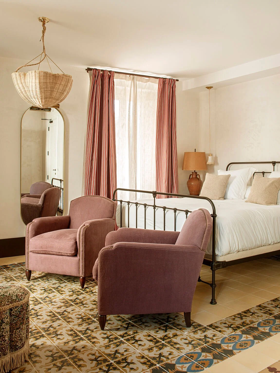 A cozy bedroom with a metal bed frame, pink armchairs, a mirror, a lamp, and patterned tile flooring.