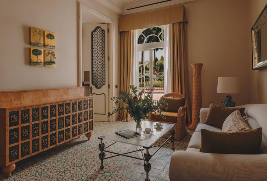 Living room with beige sofa, armchair, marble coffee table, and flowers, with large window and ornate door.