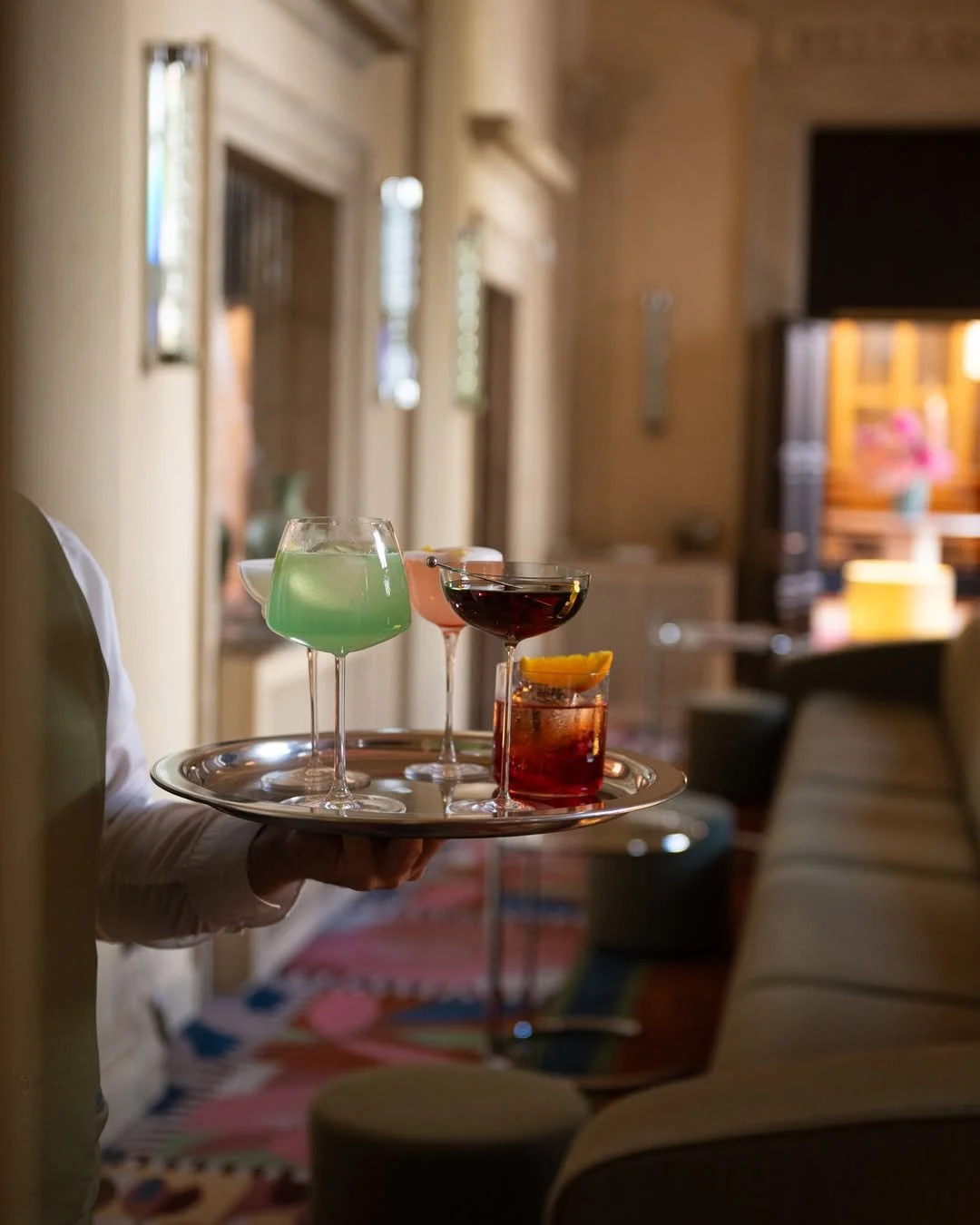 A person holding a tray with four cocktails in a cozy indoor setting, with blurred background of furniture and decor.