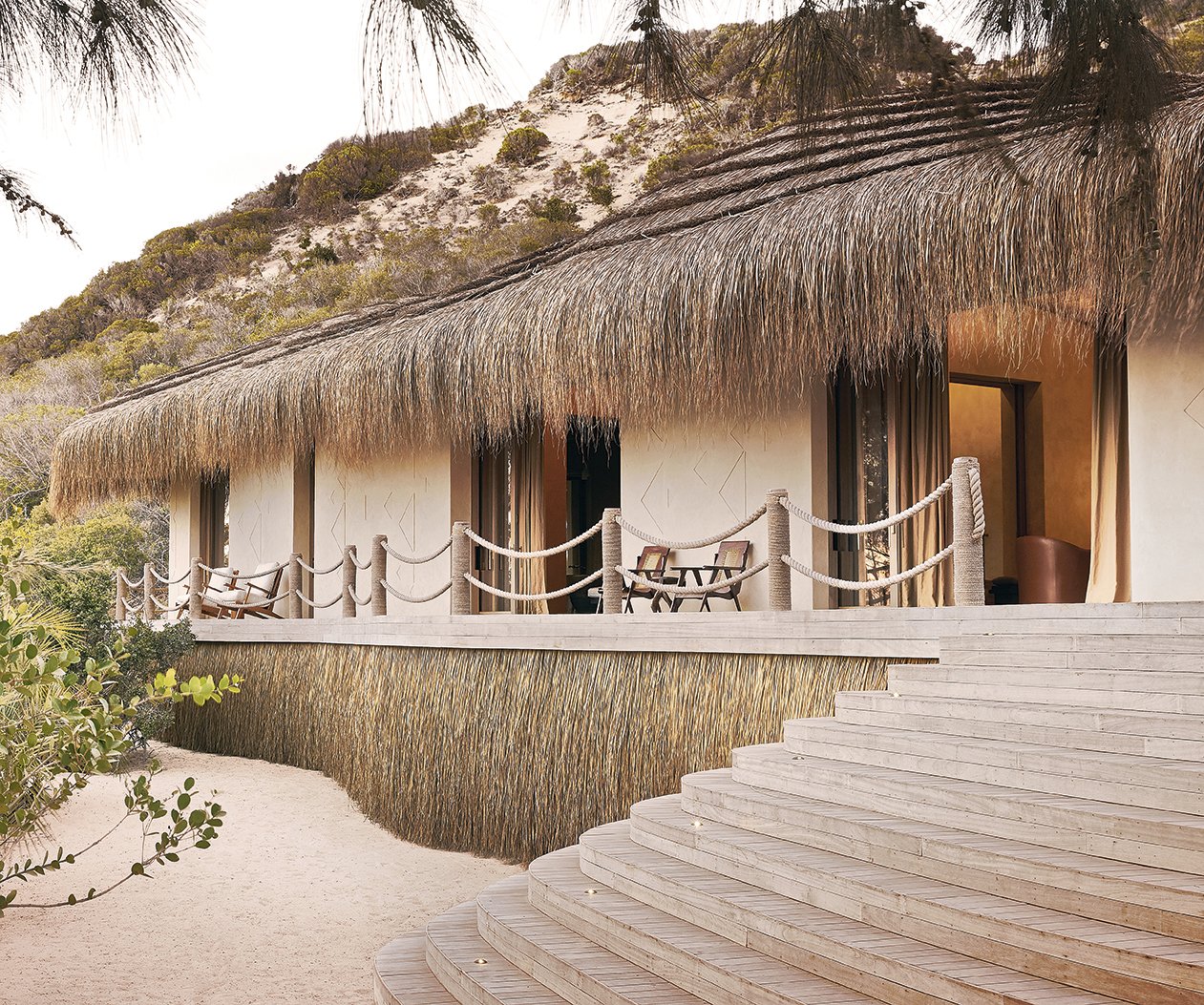 A beachfront bungalow with a thatched roof, wooden deck, and outdoor seating area, surrounded by sandy ground and lush green vegetation.