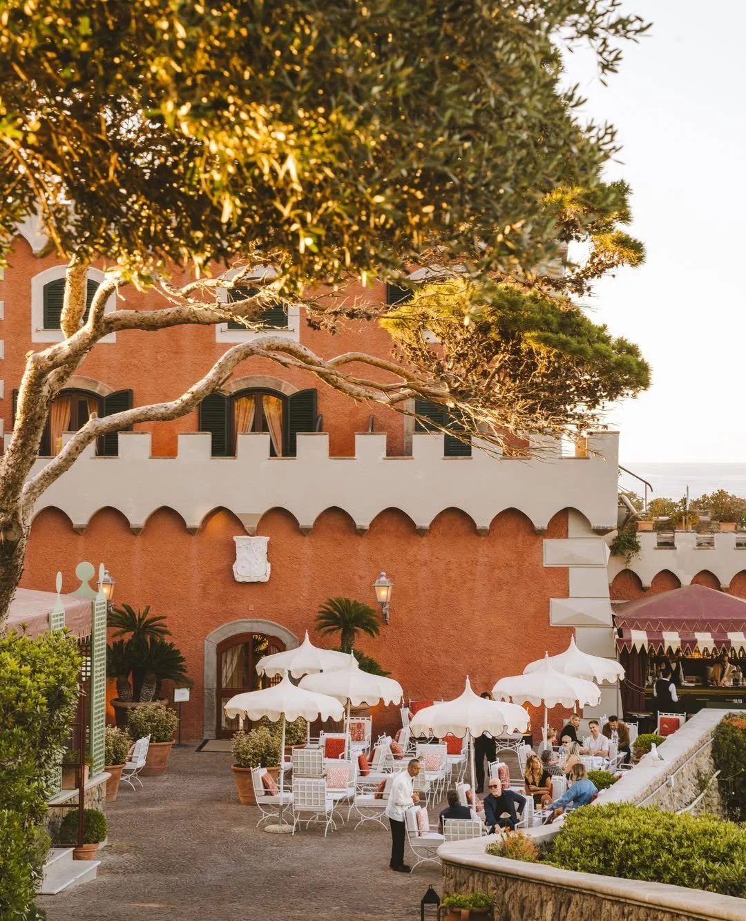 Outdoor patio of a restaurant or hotel with white tables, chairs, and umbrellas, surrounded by potted plants, with a large orange building with arched windows and a white decorative wall in the background, and people dining and socializing during sunset.