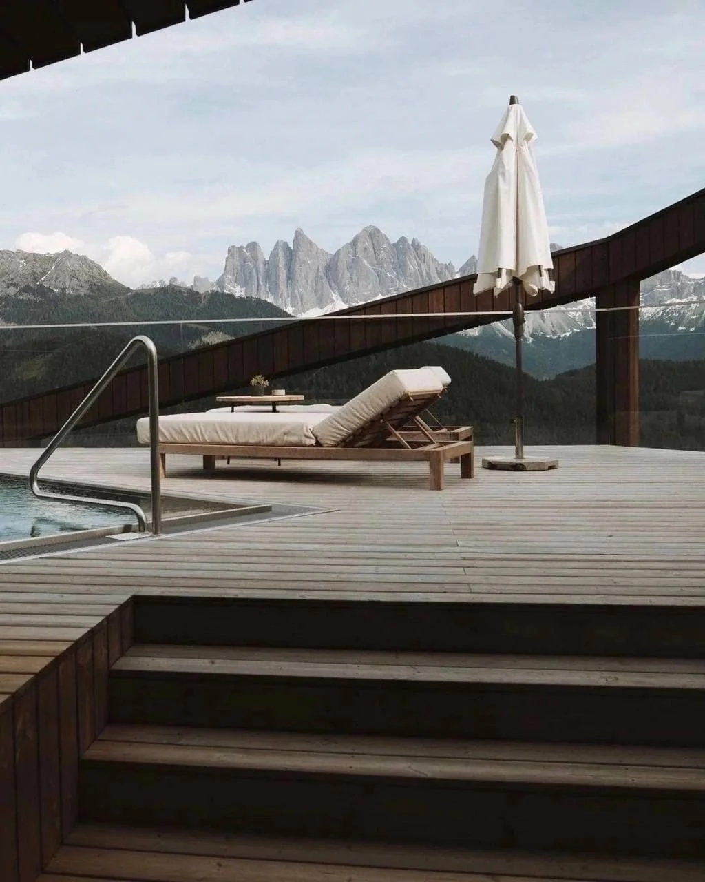 Wooden deck with pool, sunbed, table, and umbrella overlooking mountainous landscape with rocky peaks in the background.
