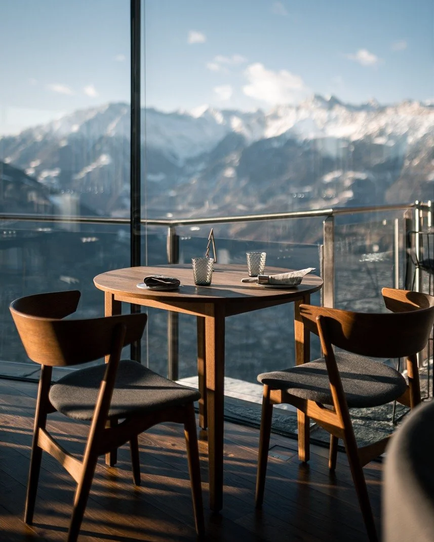 A round wooden table set with glasses and napkins on a balcony with mountain views.