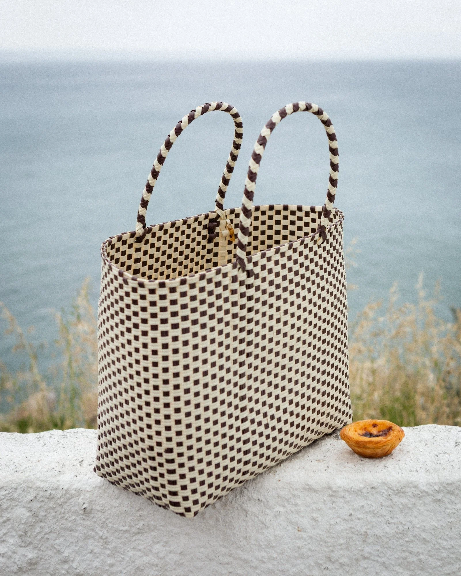 A checkered woven tote bag with dark brown and beige stripes on the handles sitting on a white surface with a small pastry nearby, against a backdrop of water and sky.