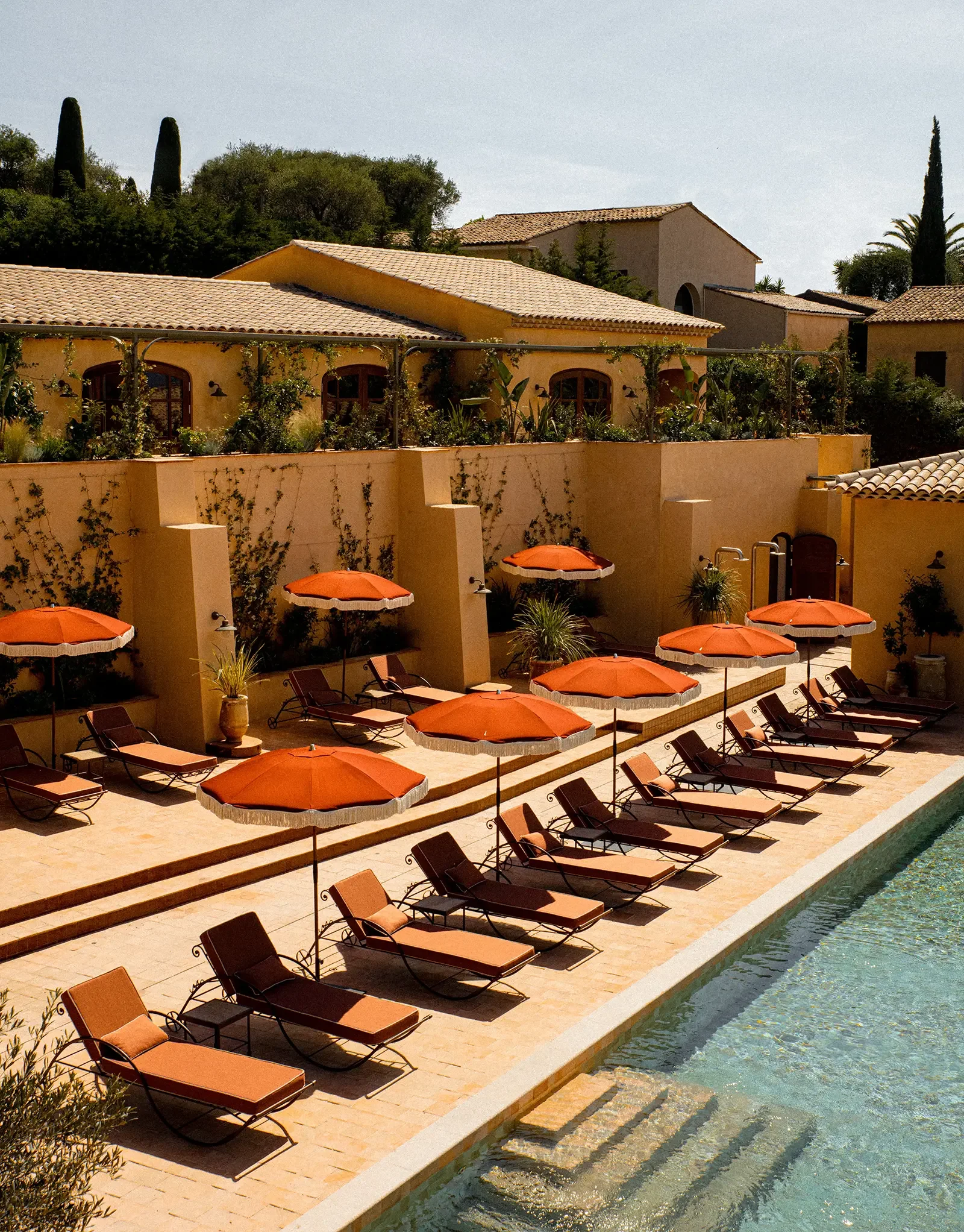 Swim pool with orange umbrellas and lounge chairs beside a beige building with arches and a garden, in a Mediterranean-style residential area with tiled roofs and tall cypress trees.