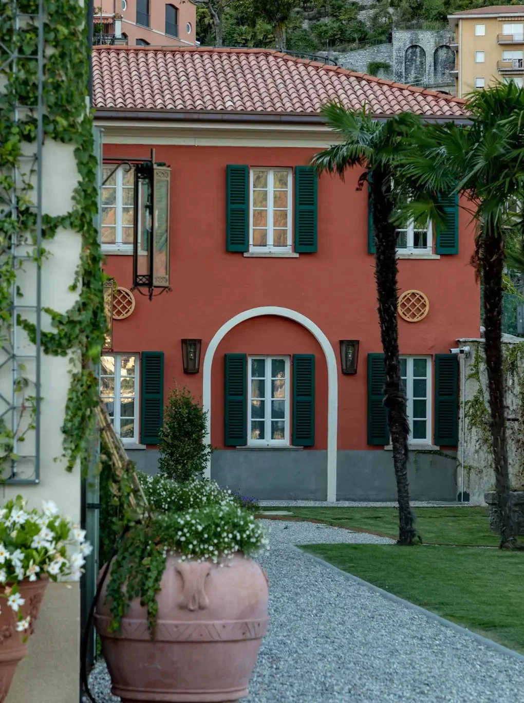 A red stucco house with green shutters, surrounded by plants and palm trees, and a gravel pathway leading to the entrance.