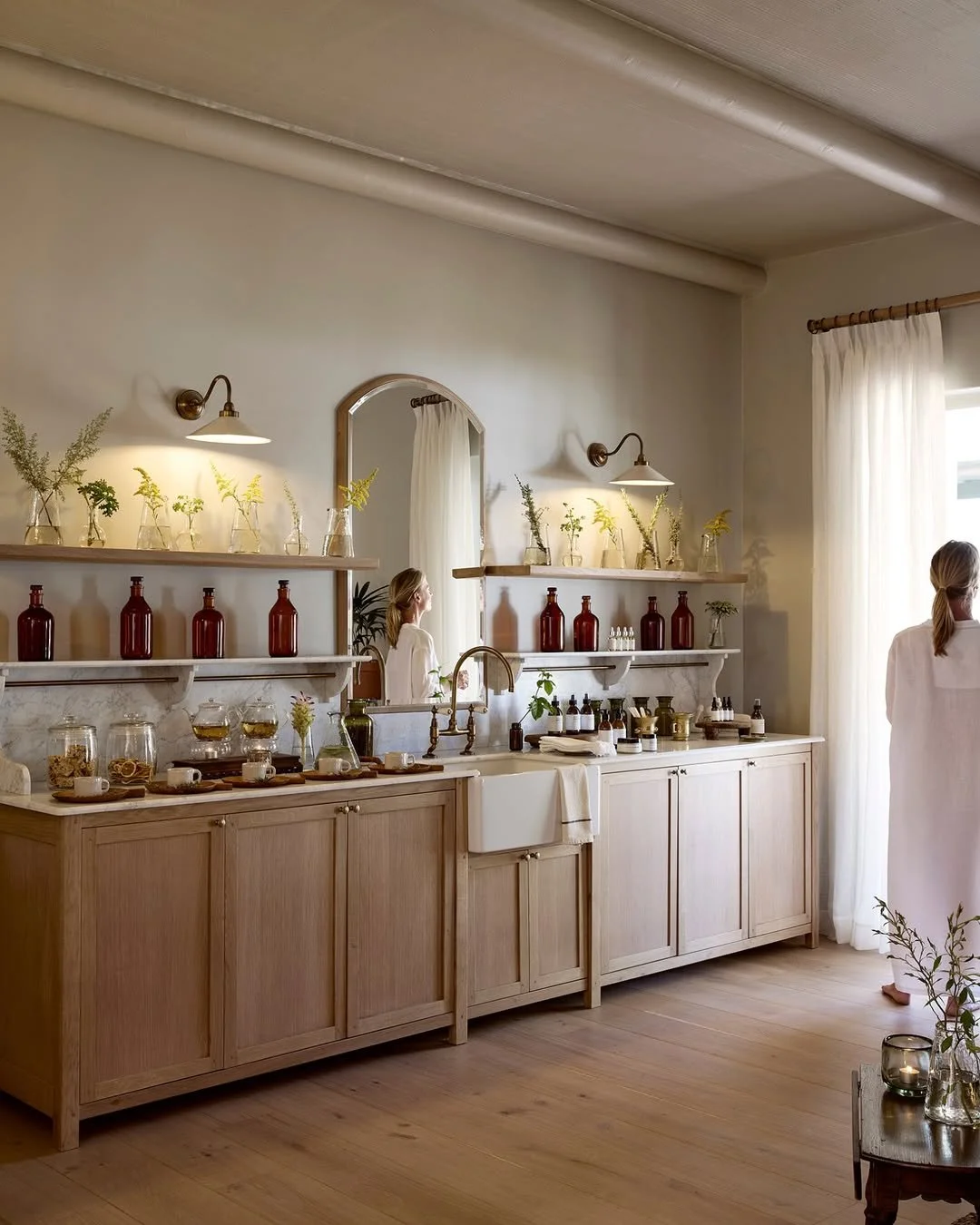 A minimalist kitchen with wooden cabinets, white countertops, and shelves with amber bottles and glass jars. A large mirror reflects a woman in a white dress, and sunlight filters through a sheer curtain.