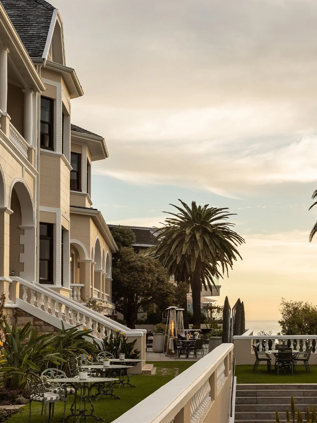 A luxurious waterfront mansion with elegant balconies, white railings, and lush landscaping including palm trees. Outdoor tables with umbrellas and seating are visible, with a view of the ocean at sunset.