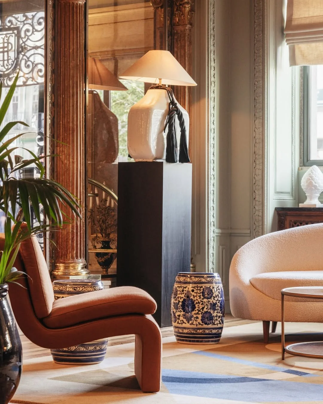 Elegant living room with vintage decor, including a large ceramic lamp on a black pedestal, plush beige chairs, decorative vases, and ornate molding along the window and wall.