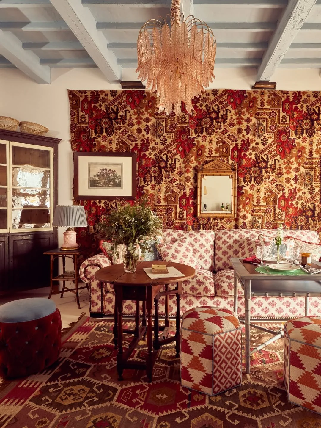 Cozy living room with a patterned red and beige tapestry on the wall, vintage furniture, a chandelier, a floral sofa, a round wooden coffee table with a vase of flowers, and patterned ottomans.