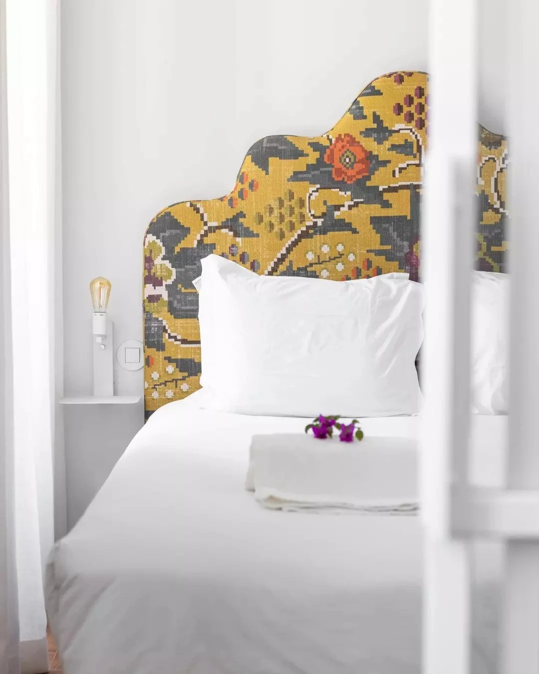 A neatly made bed with white sheets and pillows, set against a distinctive floral patterned headboard. A small bedside table holds a modern lamp with an exposed lightbulb and a flower arrangement.