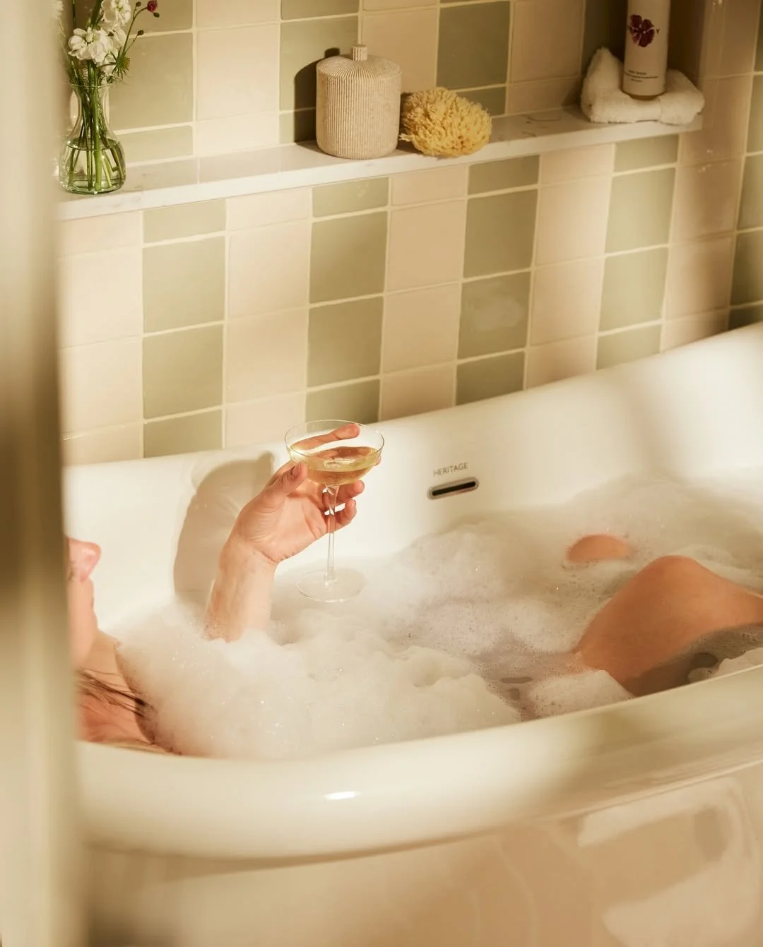 Person relaxing in a bubble bath holding a glass of white wine with various bathroom items on a tiled shelf behind them.