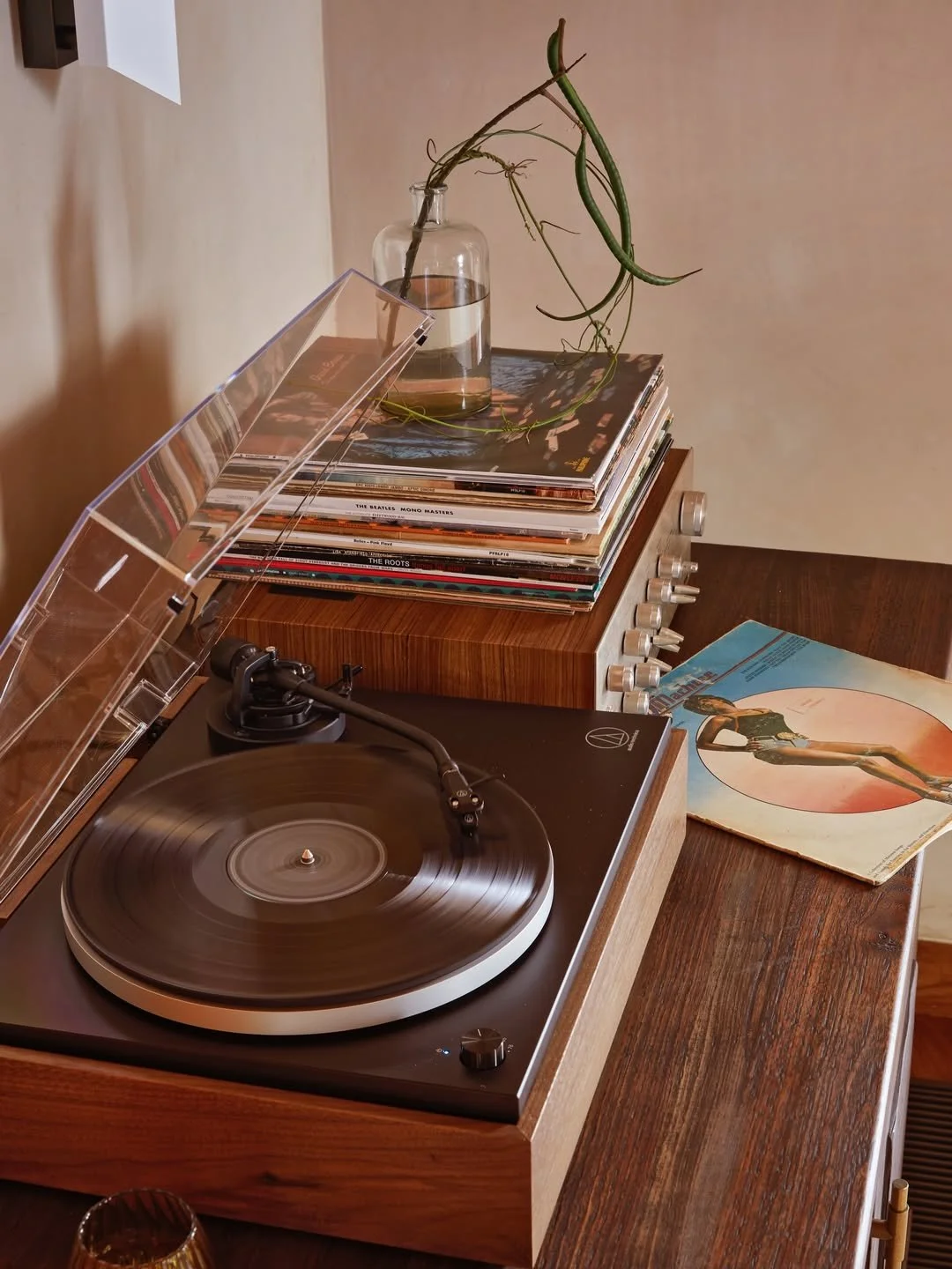 A vintage turntable on a wooden surface with a vinyl record spinning, a stack of vinyl records nearby, a vinyl record cover with an artistic illustration, a glass vase with a plant, and a collection of books or magazines on top of a vintage amplifier.