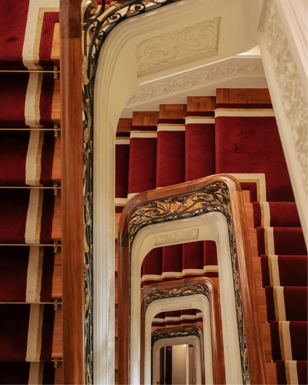 Overhead view of a spiral staircase with red carpeted steps, ornate iron railings, wooden handrails, and decorative wall moldings.