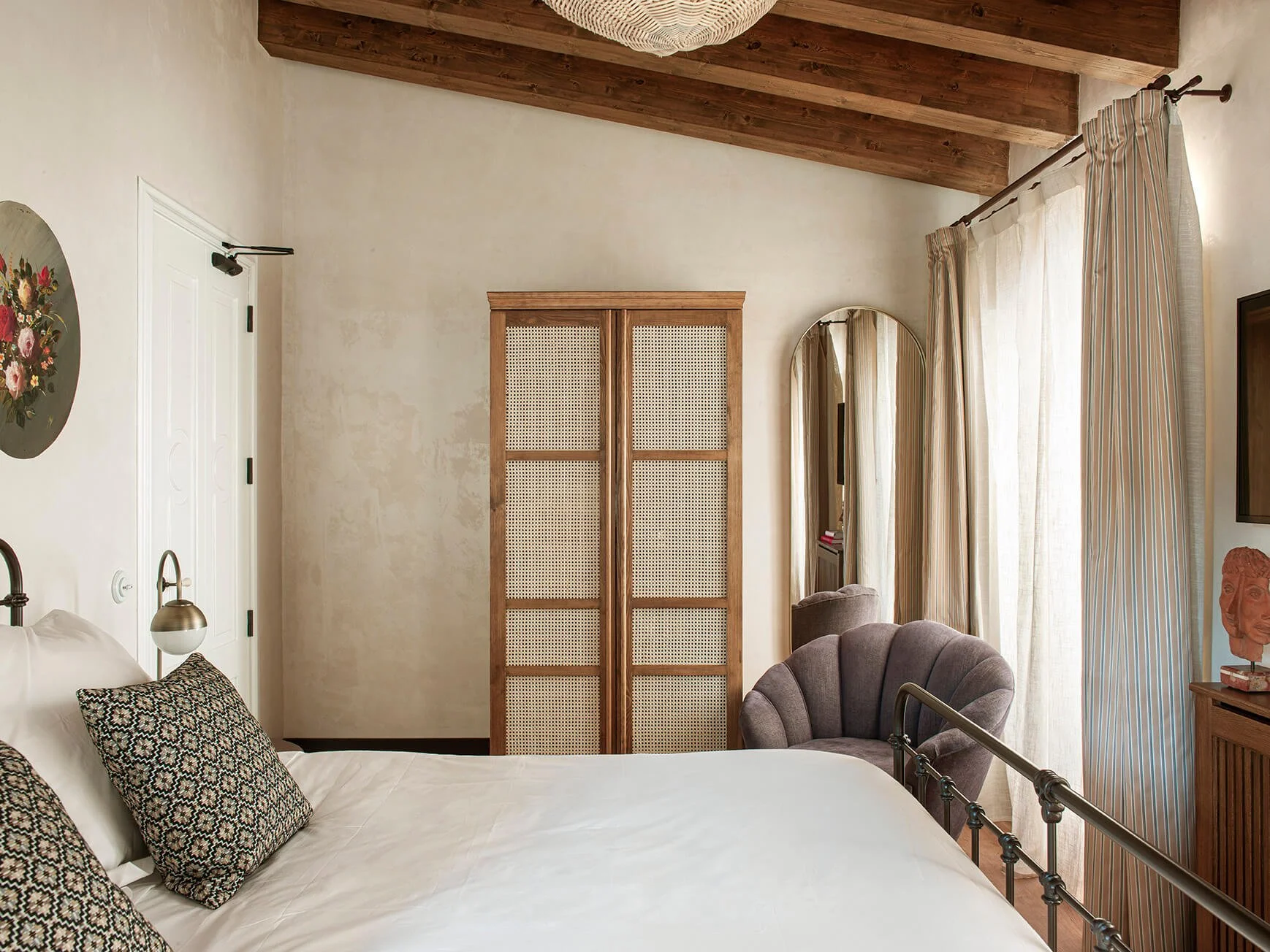 A bedroom with a bed, pillows, a wall-mounted lamp, a wooden wardrobe with woven doors, a mirror, a window with striped curtains, a gray armchair, and a wooden table with a bust sculpture.