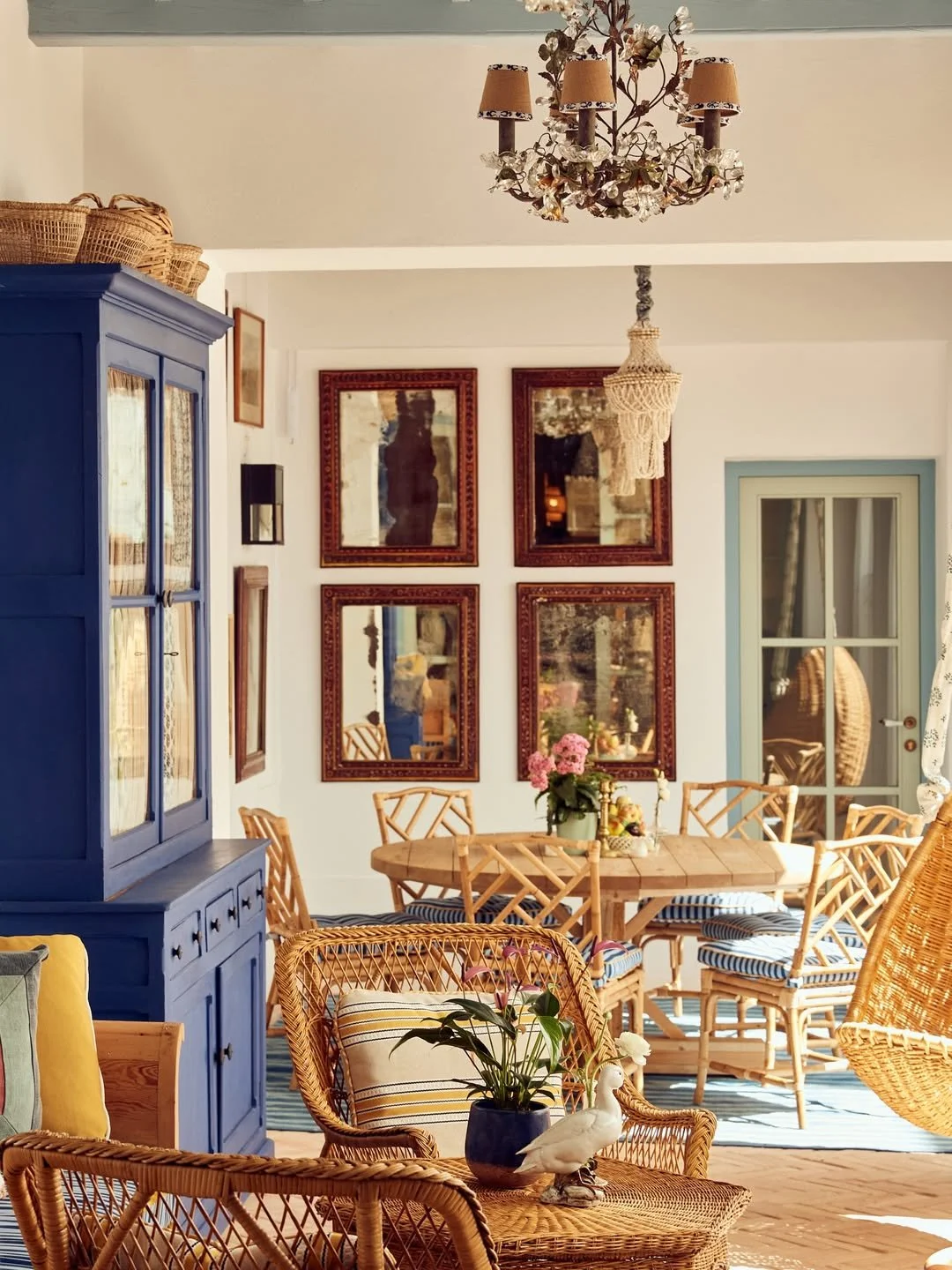 A cozy dining area with wicker chairs, a blue cabinet, framed mirrors on the wall, a chandelier, a table with a flower arrangement, and a glass door leading outside, decorated in a rustic style.