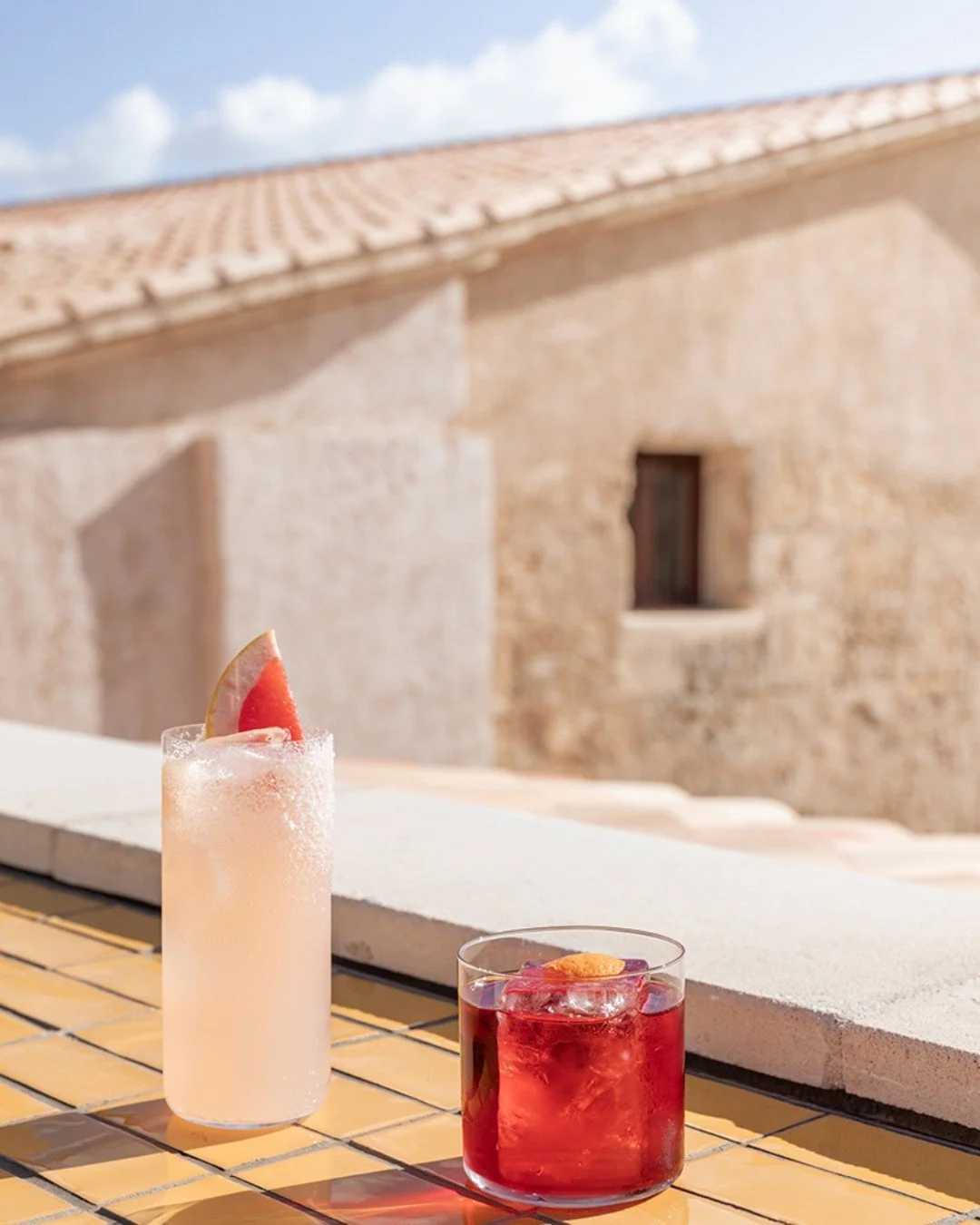 Two cocktails, one pink with a slice of watermelon garnished on top, and one red with an orange peel, sit on a terracotta tiled surface outdoors with a stone building and a partly cloudy sky in the background.