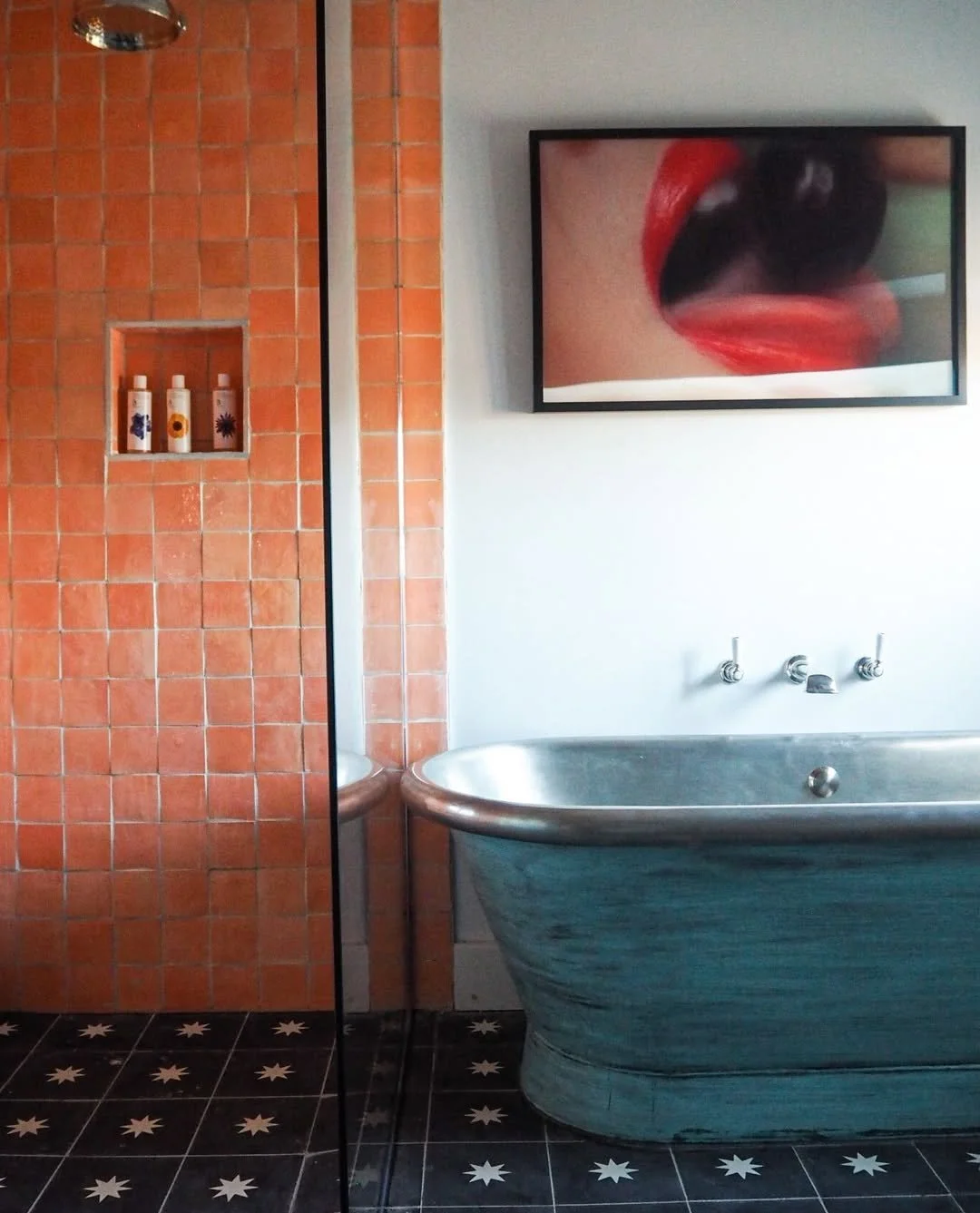 A bathroom featuring a vintage-style blue bathtub, a black framed artwork of a close-up of lips with red lipstick, partially visible black and white star-patterned floor tiles, and a shower area with orange tiles and bottles on a shelf.