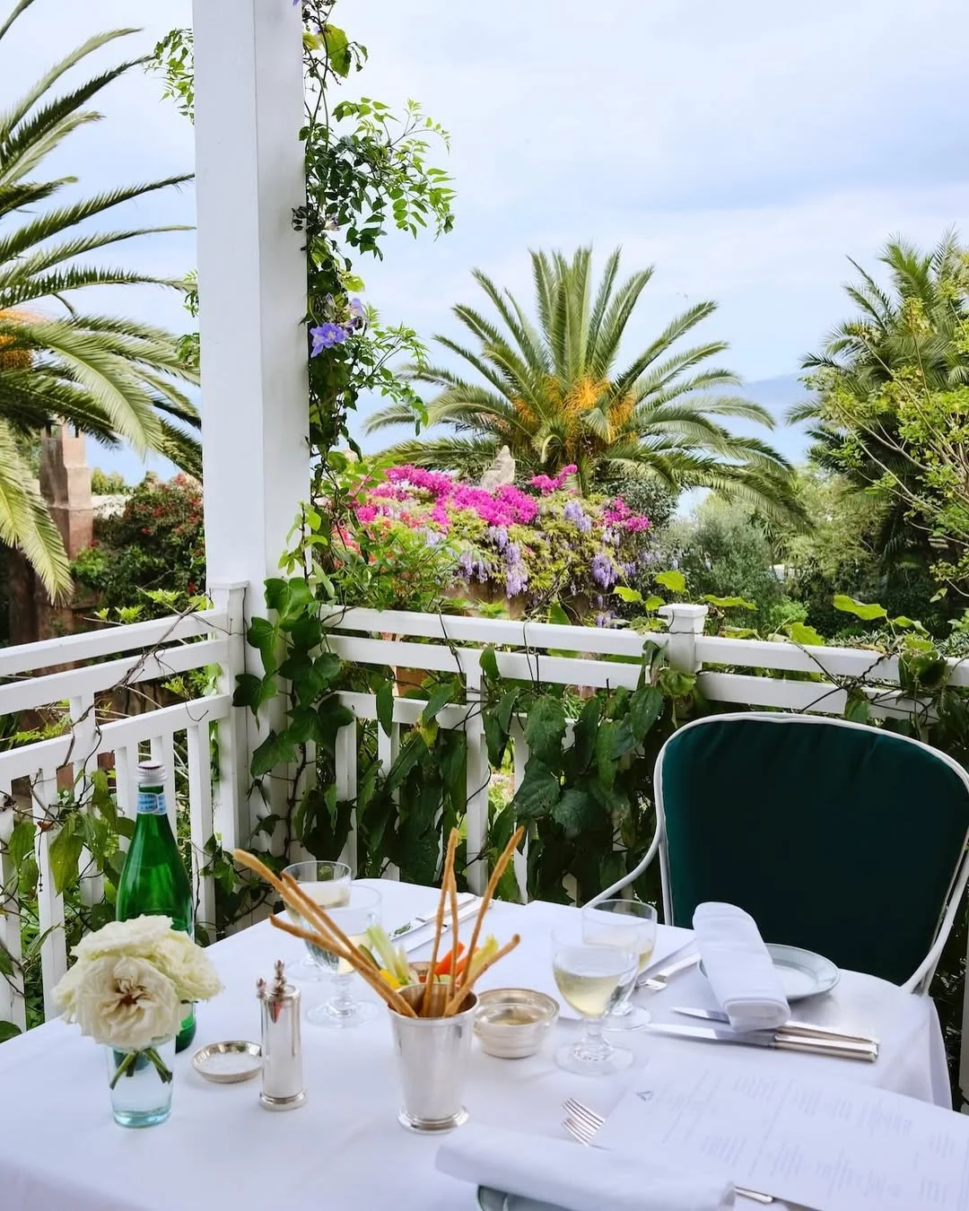 A table set for dining on a porch with lush greenery and colorful flowers in the background, including palm trees and blooming plants.