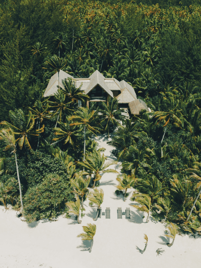 Aerial view of a tropical beach with white sand, surrounded by dense green palm trees and a large building with a gray roof in the background.