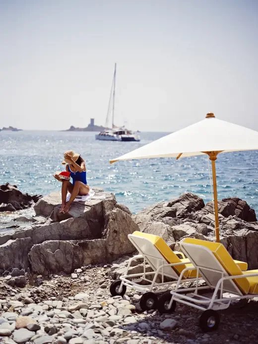 A woman in a blue swimsuit and large sunhat sitting on rocks by the beach, with two yellow lounge chairs and a white umbrella nearby, while sailboats are in the water and a lighthouse is visible in the distance.