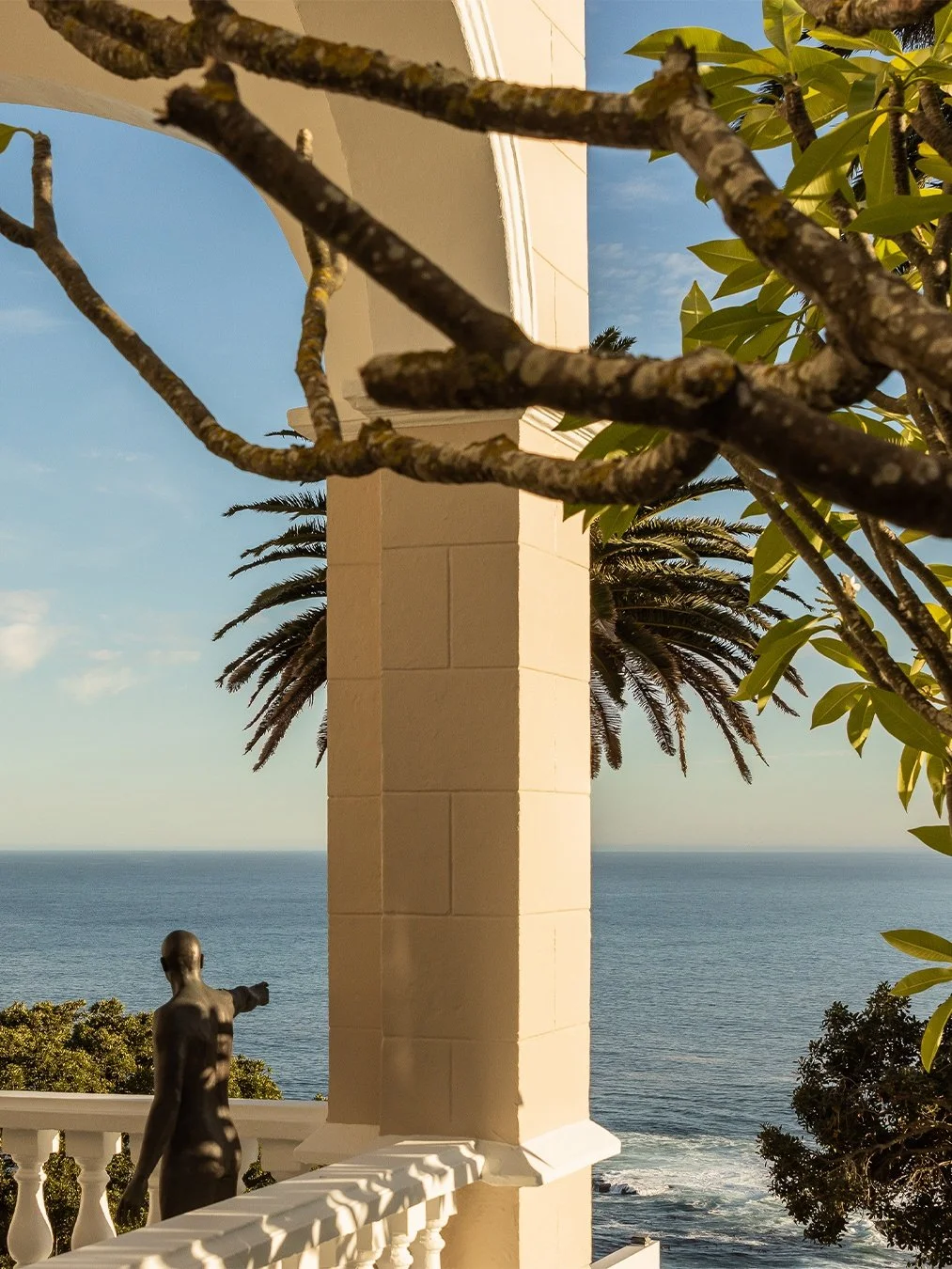 View of the ocean from a balcony with a cream-colored pillar, a statue of a woman pointing, and tropical trees including palm and other leafy trees.