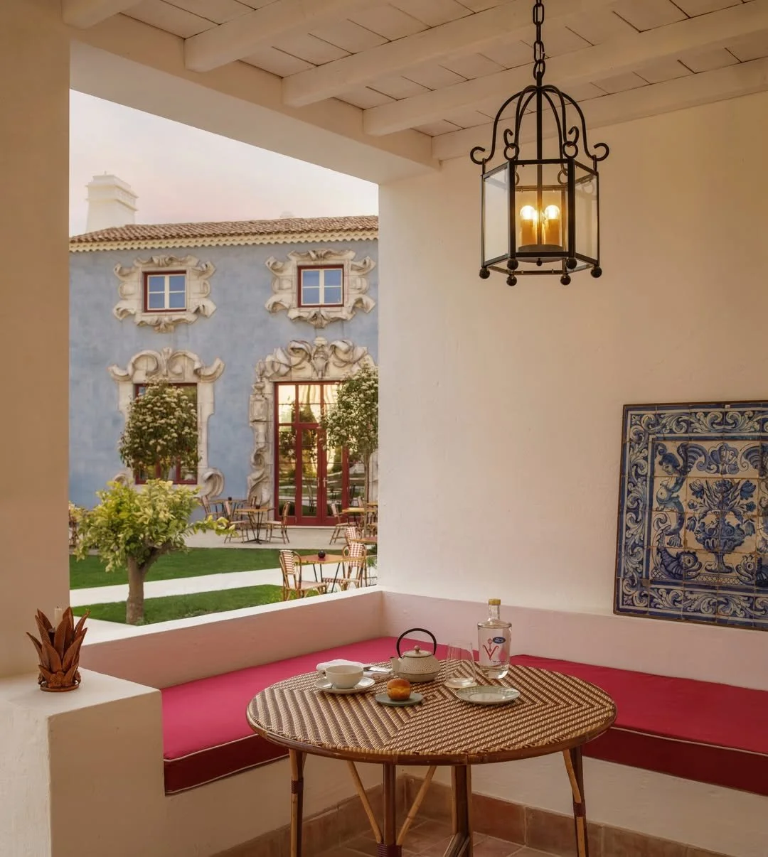A cozy porch with a round wicker table set for tea, featuring a teapot, cup, and bottle. A pink cushioned window seat runs along the white wall. A decorative blue tile hangs on the wall, and a black wrought iron lantern hangs from the ceiling. Outside, a courtyard with small trees, outdoor tables, and a pastel blue building with ornate white window frames and a clay tiled roof is visible.