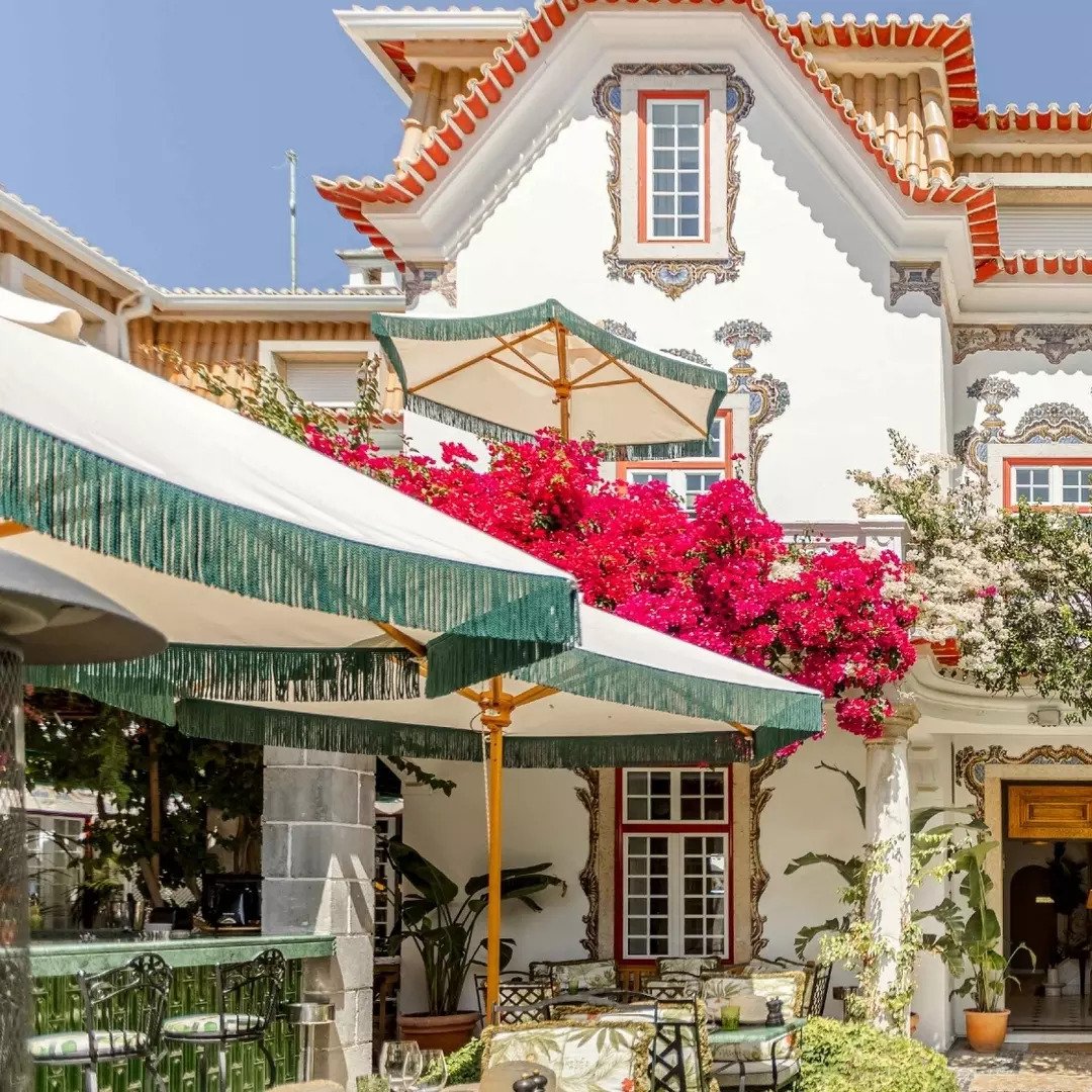 A white house with decorative trim, pink and white flowering trees, green and white striped umbrellas, outdoor seating, and potted plants, under a clear blue sky.