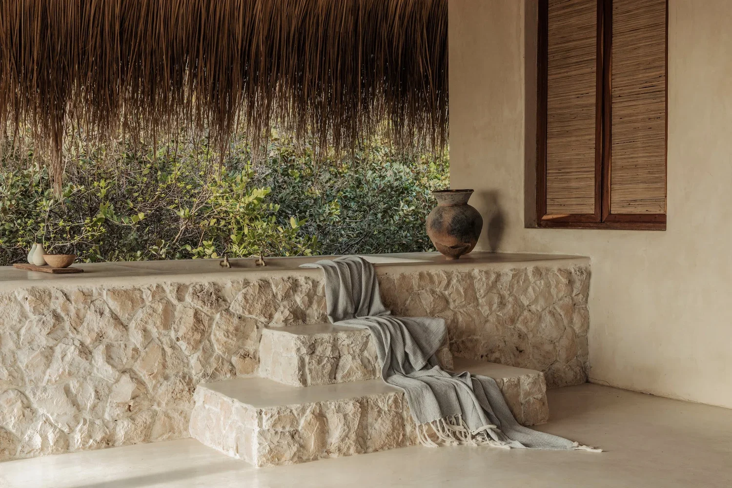 A stone staircase with a gray blanket draped over it, set against a textured stone wall wall with a large clay vase and a wooden window with closed shutters.