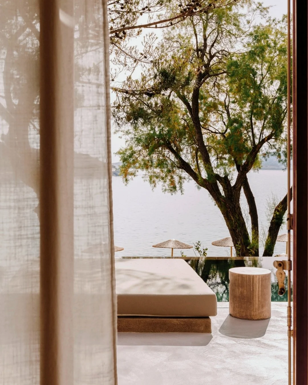 View of a tree and lake seen from a patio with a lounge chair and two straw umbrellas in the background, partially obscured by semi-transparent curtain.
