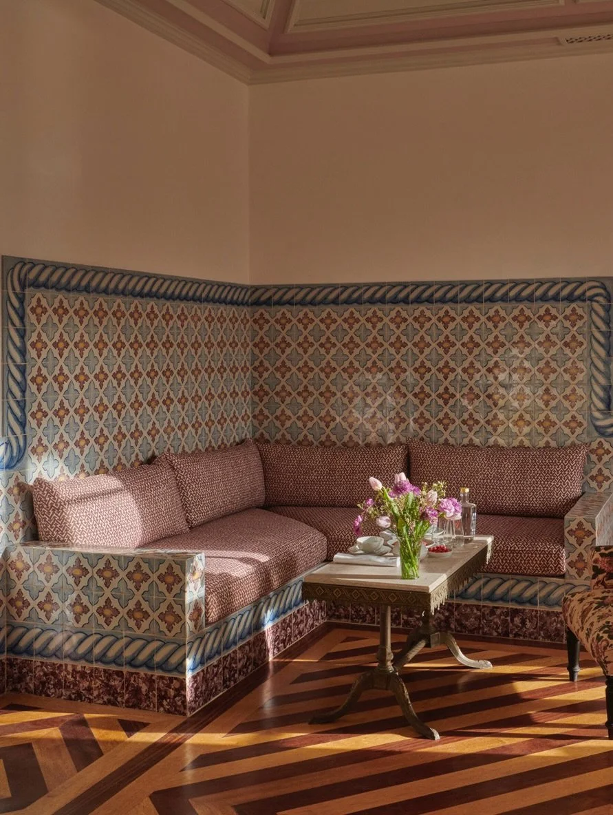 A corner of a room with a built-in tiled bench with red-patterned cushions, a small wooden table with a flower vase, glasses, and plates, and a floral armchair; parquet wood floor and textured wall with a decorative ceiling.