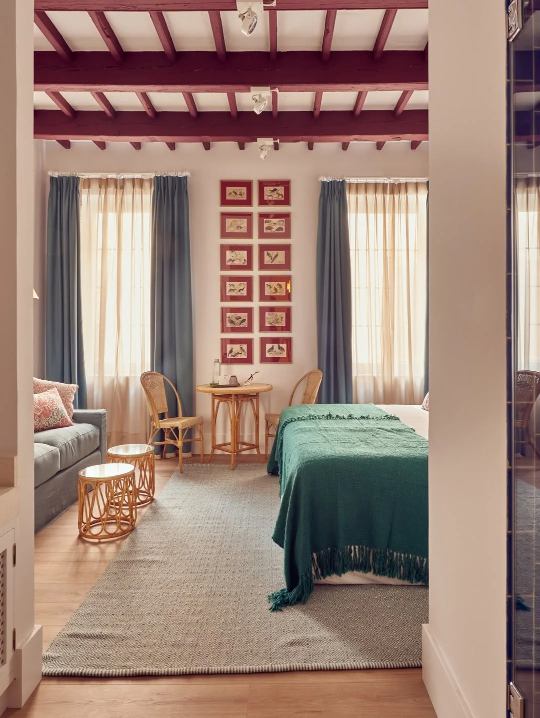 Interior of a cozy bedroom with a wooden beamed ceiling, beige walls, and two large windows with beige and blue curtains. A small round table with two chairs is against the wall, decorated with framed artwork of animals arranged vertically above it. A bed covered with a green blanket is on the right, and a light gray sofa with patterned pillows is on the left, with two small matching side tables beside it. The room has a neutral-toned area rug and natural light streaming in.