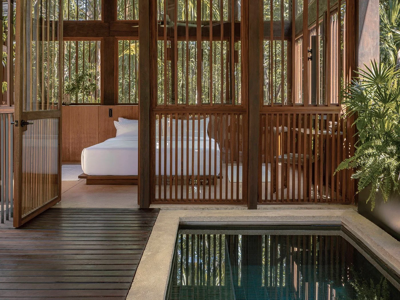 Bedroom with white bed inside a wooden enclosed patio, adjacent to a small pool, with large glass doors and tropical plants.