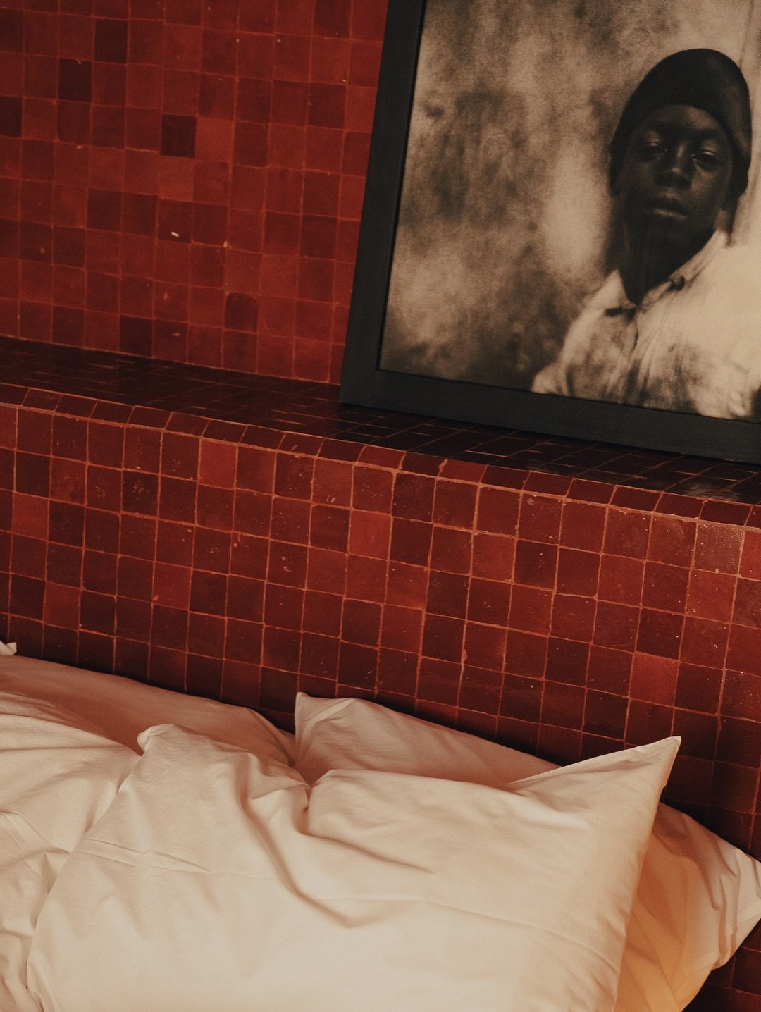 A bed with white sheets and pillows in front of a red tiled wall. A framed black and white portrait of a person with dark skin, wearing a turban and a light shirt, hangs above the headboard.