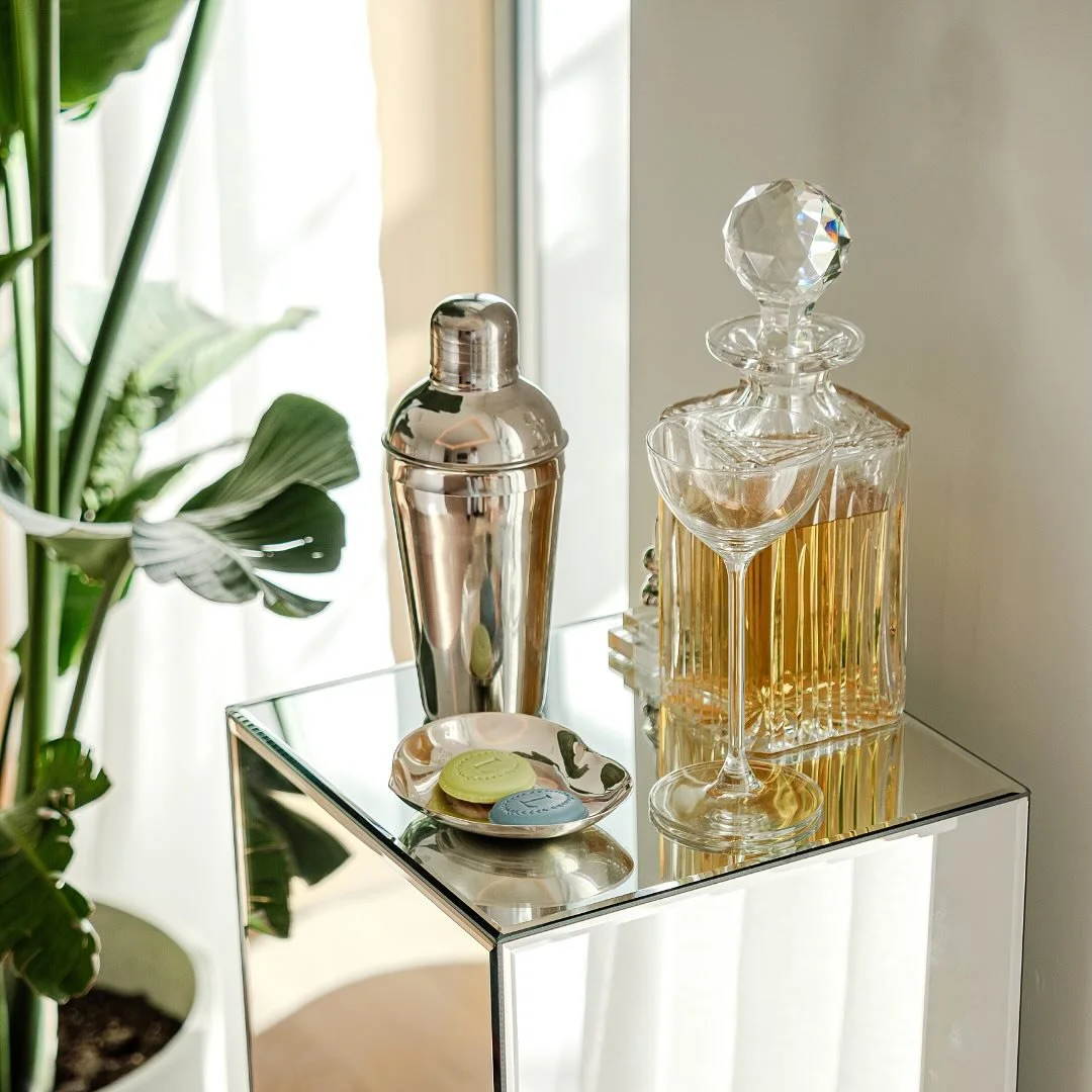 A mirrored side table holding a cocktail shaker, decanters, two cocktail glasses, and colored coasters, with a large green plant nearby.