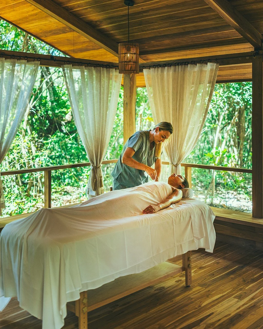 A woman lying on her back on a massage table outdoors, receiving a massage from a therapist in a wooden room with open walls and white curtains, surrounded by lush green trees.