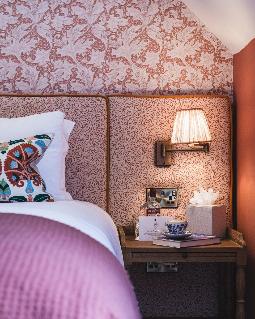 Close-up of a cozy hotel bedroom corner featuring a wooden nightstand with a tissue box, a teacup and saucer, a bottle of liquor, and a white card. A colorful embroidered pillow pattern is on the bed, with pink patterned wallpaper and a wall-mounted lamp with a striped shade illuminating the area.